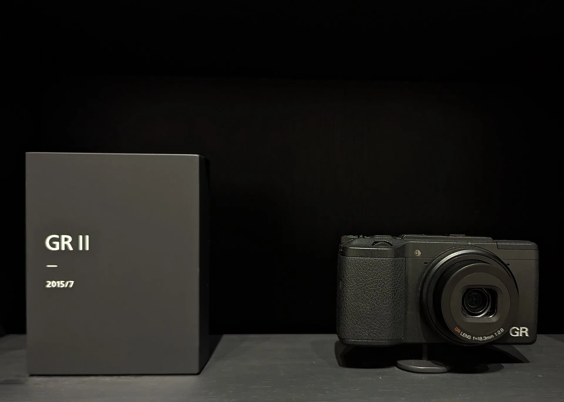 A black Ricoh GR II on a plinth beside a label plate reading "GR II, 2015/7", against a dark background. The lens is marked 18.3mm f/2.8.