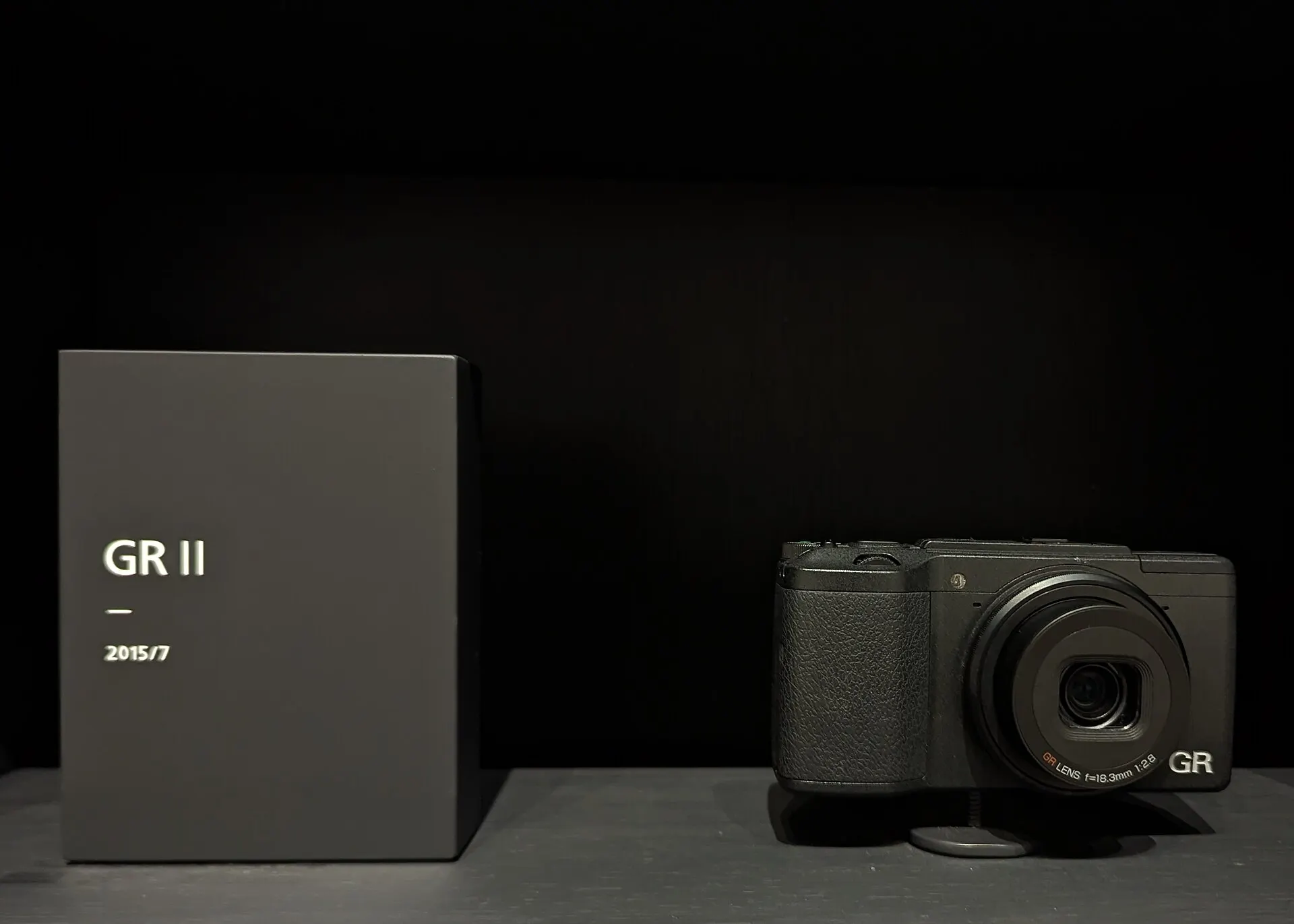 A black Ricoh GR II on a plinth beside a label plate reading "GR II, 2015/7", against a dark background. The lens is marked 18.3mm f/2.8.