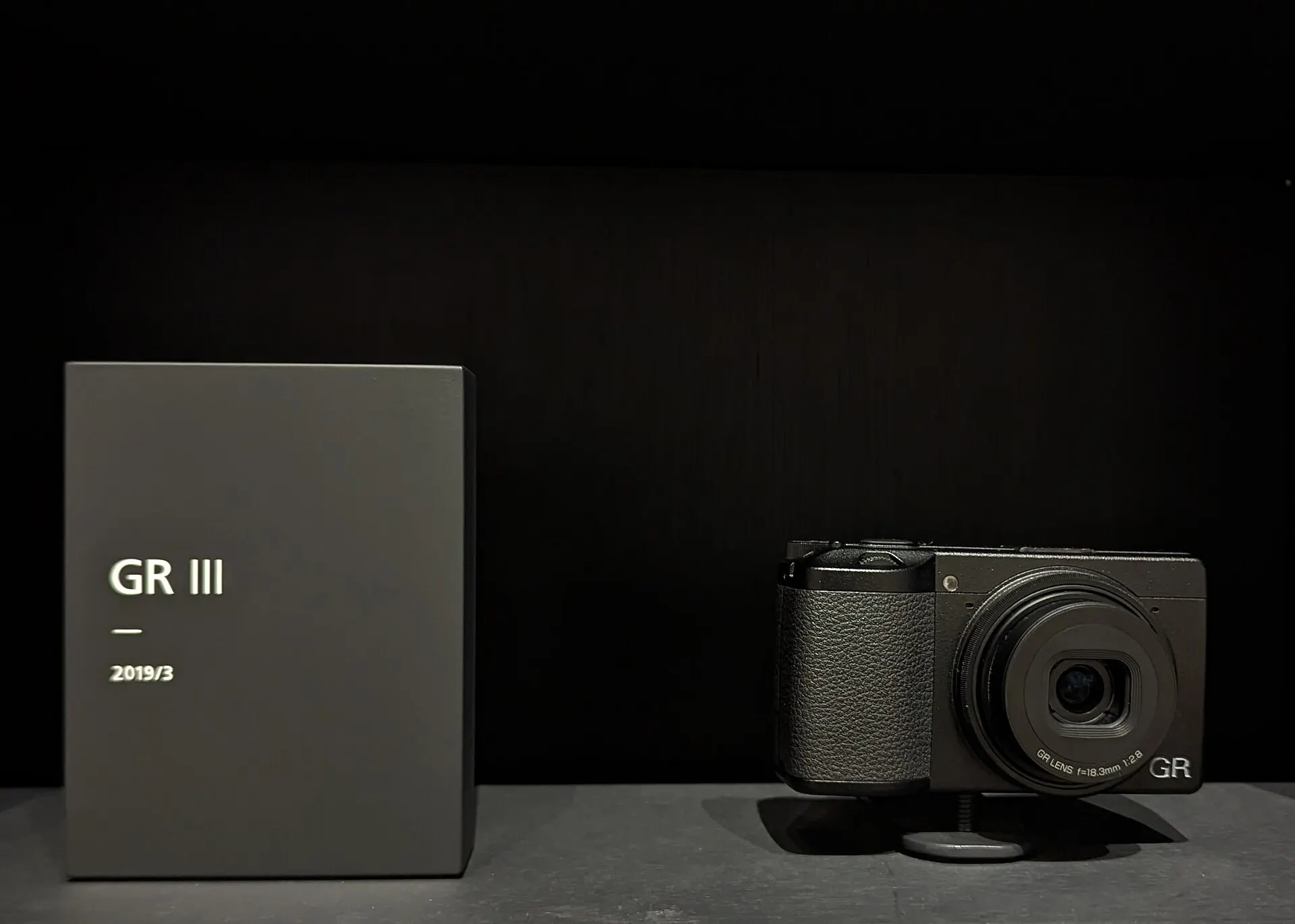 A black Ricoh GR III on a plinth beside a label plate reading "GR III, 2019/3", against a dark background. The lens is marked 18.3mm f/2.8.