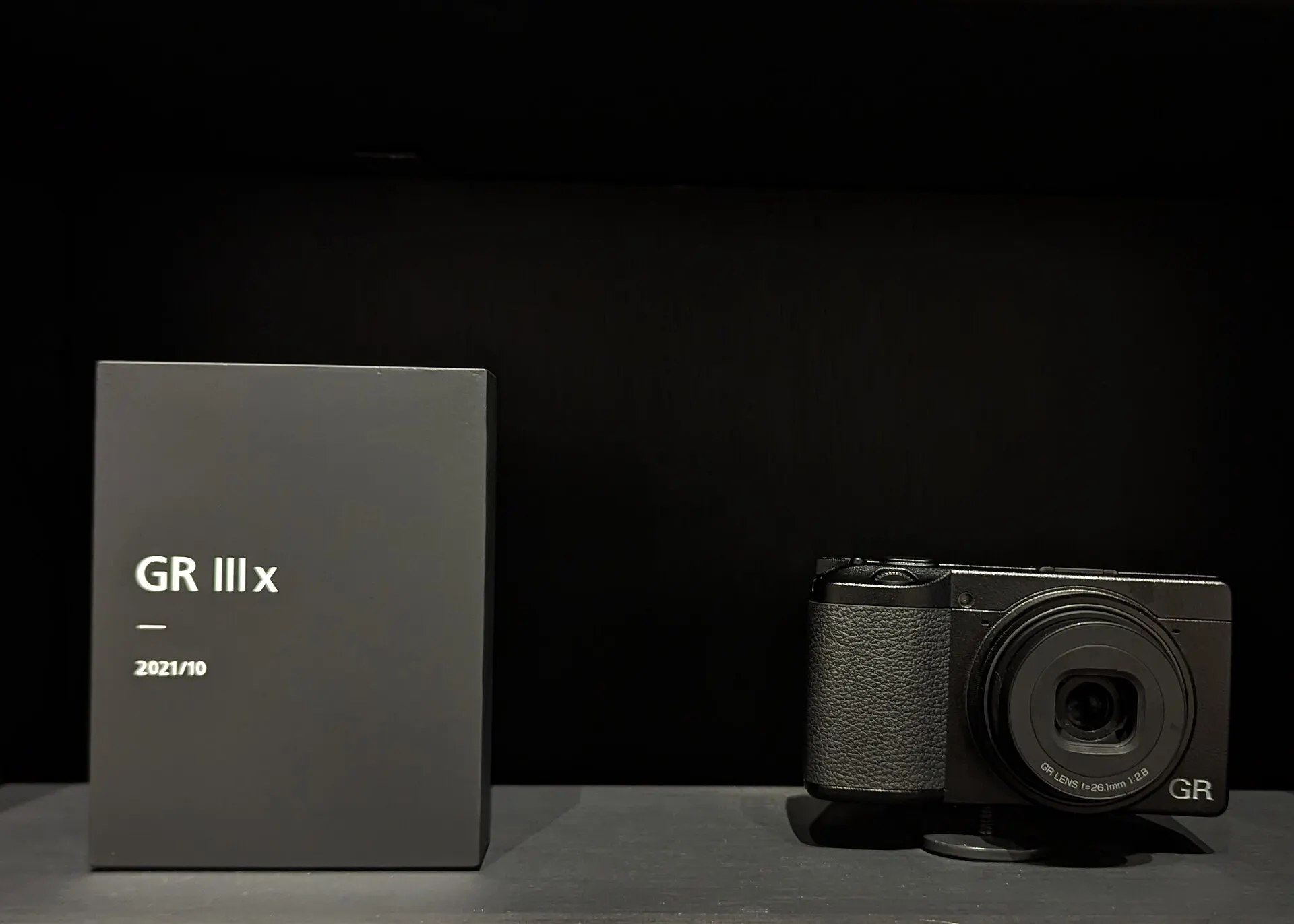 A black Ricoh GR IIIx on a plinth beside a label plate reading "GR IIIx, 2021/10", against a dark background. The lens is marked 26.1mm f/2.8.
