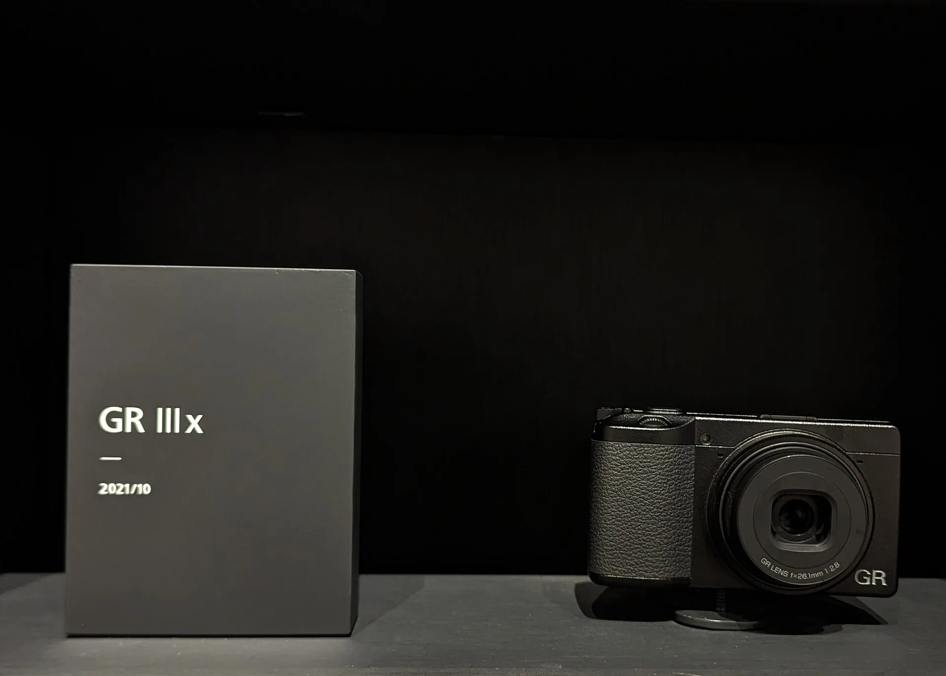 A black Ricoh GR IIIx on a plinth beside a label plate reading "GR IIIx, 2021/10", against a dark background. The lens is marked 26.1mm f/2.8.