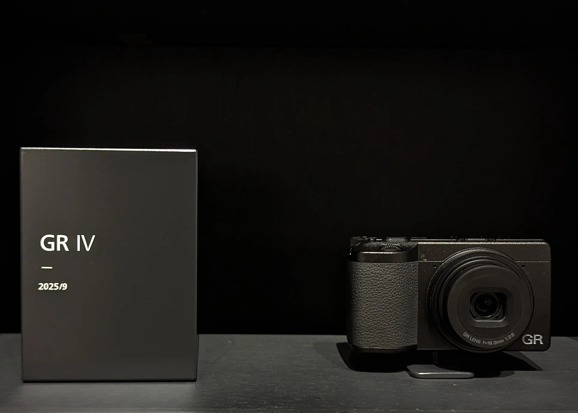 A black Ricoh GR IV on a plinth beside a label plate reading "GR IV, 2025/9", against a dark background. The lens is marked 18.3mm f/2.8.