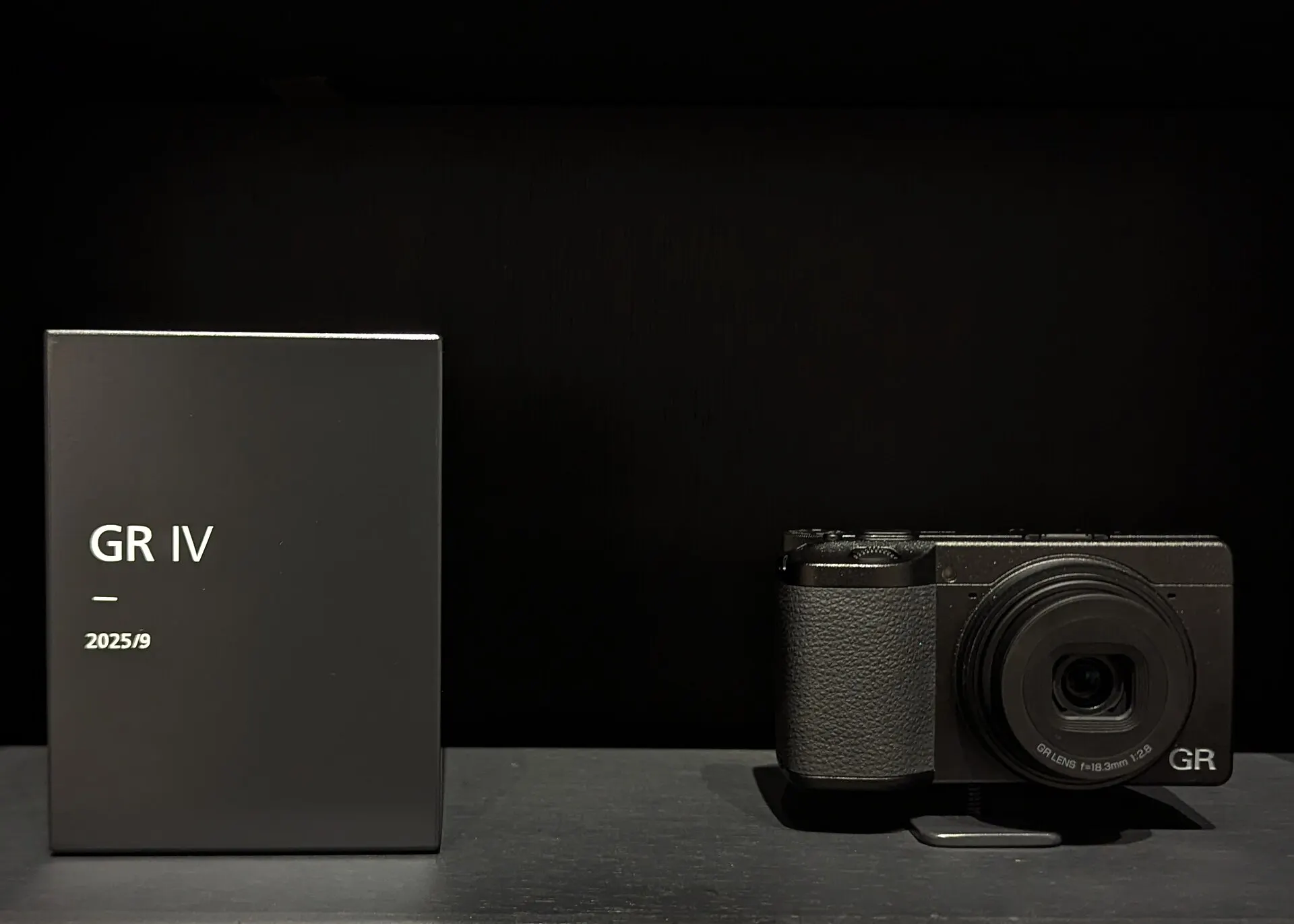 A black Ricoh GR IV on a plinth beside a label plate reading "GR IV, 2025/9", against a dark background. The lens is marked 18.3mm f/2.8.